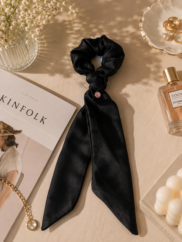 Gala black silk scrunchie with ribbon tails and pink crystal — editorial flat lay